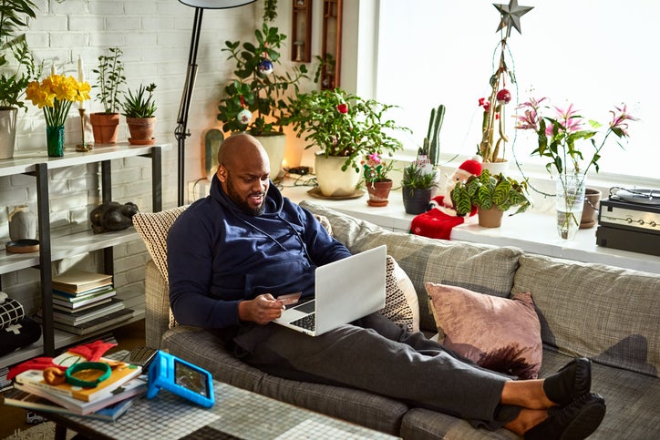 A man sitting on his couch surrounded by plants in a sunny room while online shopping on his laptop and holding a credit card in one hand.