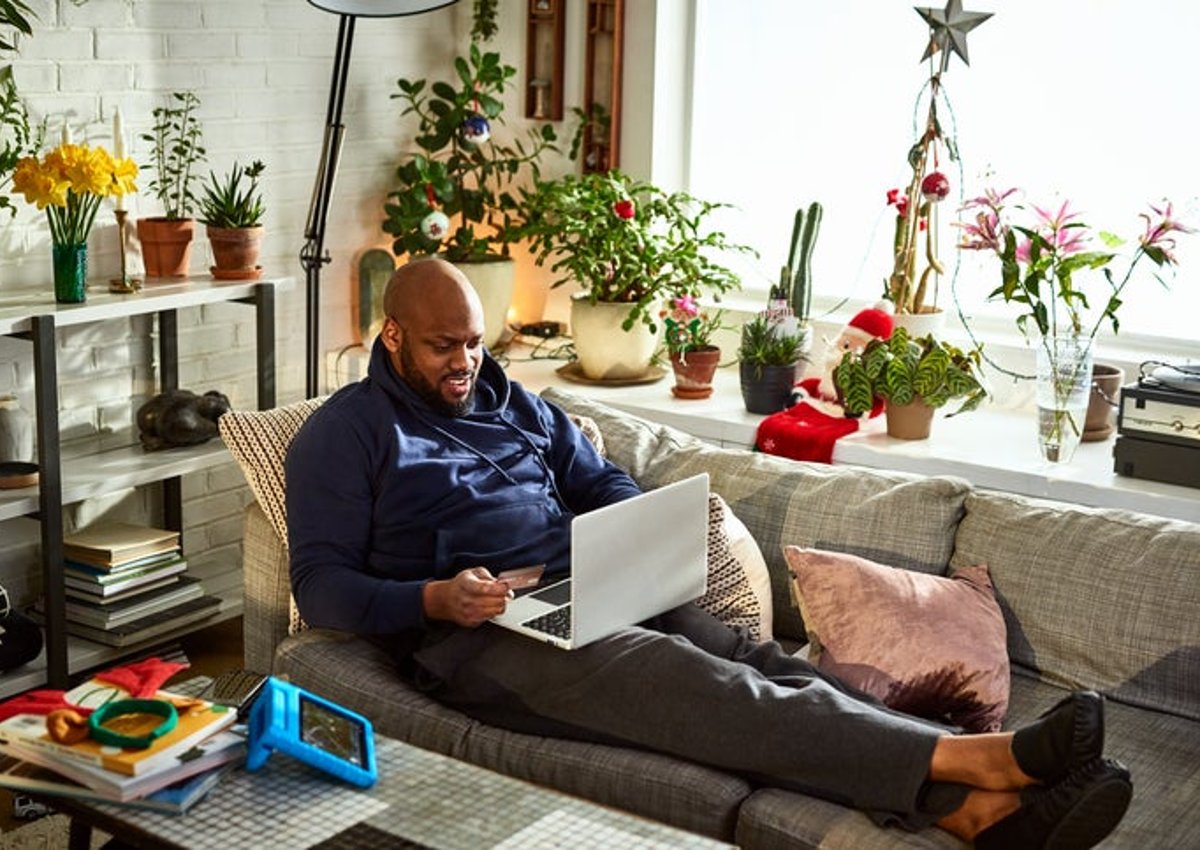 A man sitting on his couch surrounded by plants in a sunny room while online shopping on his laptop and holding a credit card in one hand.