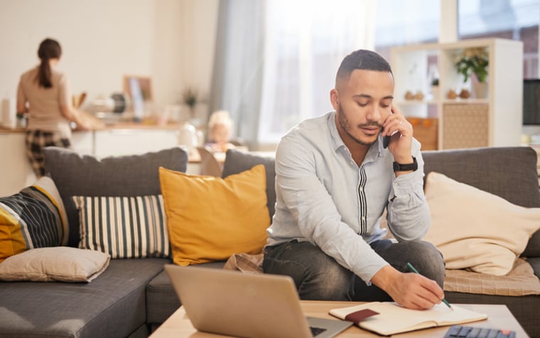 A man sitting on a couch in his living room and writing in a notebook while on the phone, with a laptop and calculator on the coffee table in front of him.