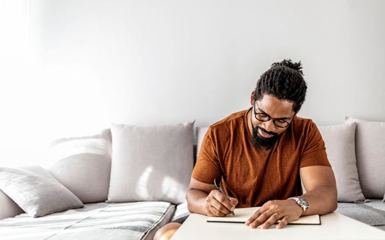 A man sat on his couch in his sunny living room and wrote in a notebook.