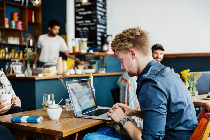 A man is typing on a laptop while sitting at a table in a cafe.