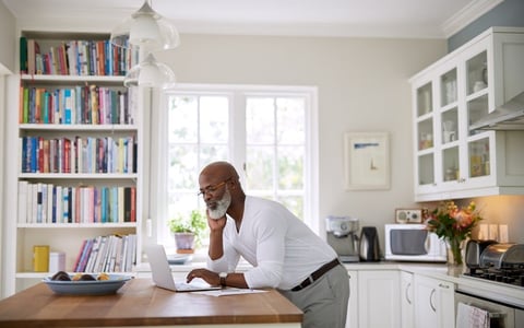 A man standing at the counter in his sunny kitchen and typing on his laptop.