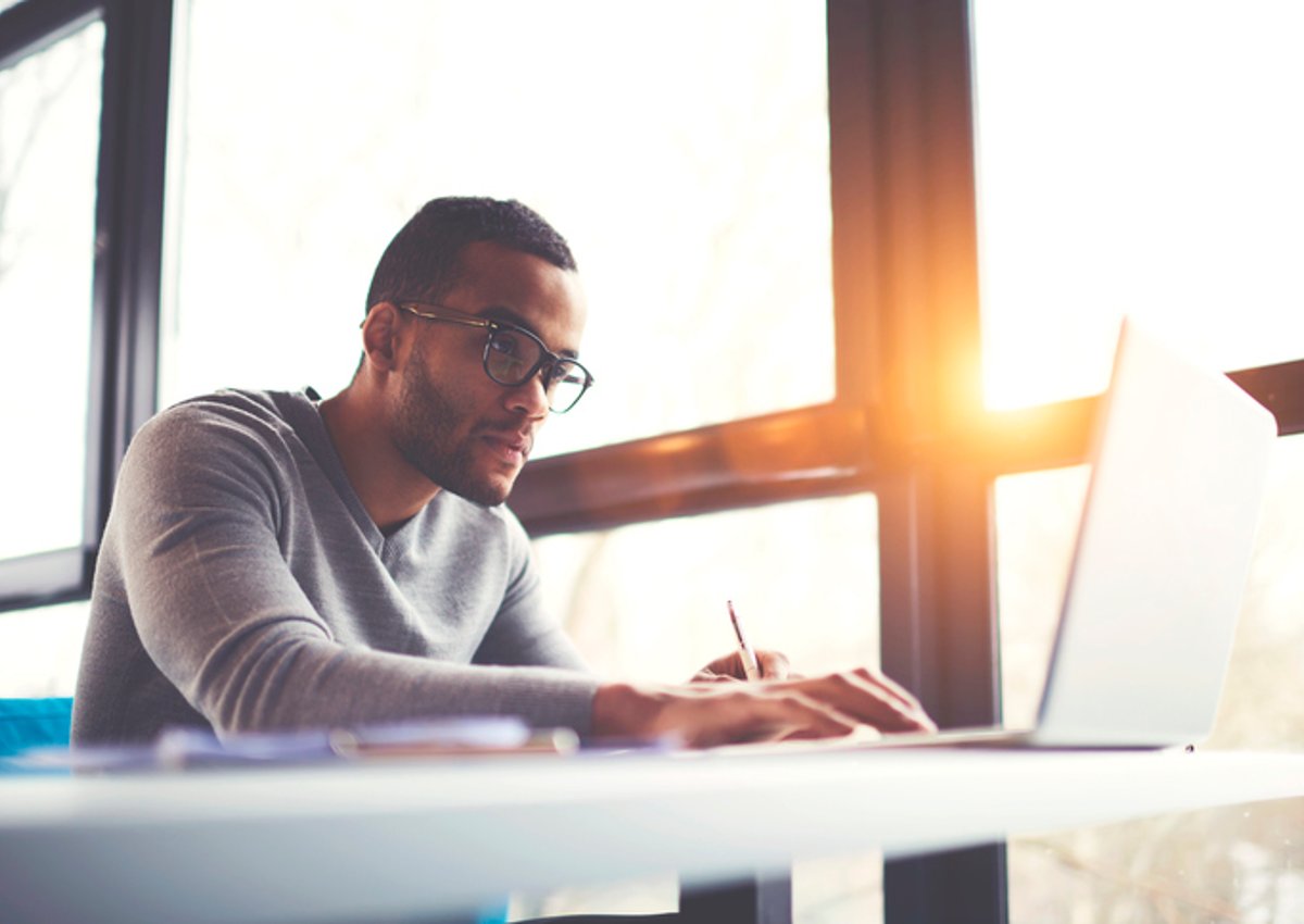 A man concentrating on his laptop while sitting next to a sunny window.