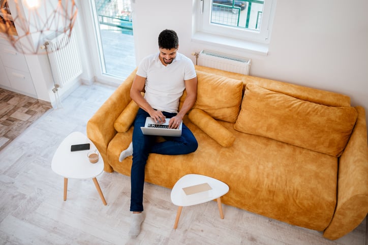 A man sitting on a couch in a sunny living room and typing on his laptop.