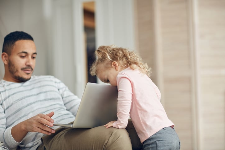 A man sitting on the couch and looking at his laptop while his toddler daughter peeks over the top of the screen.