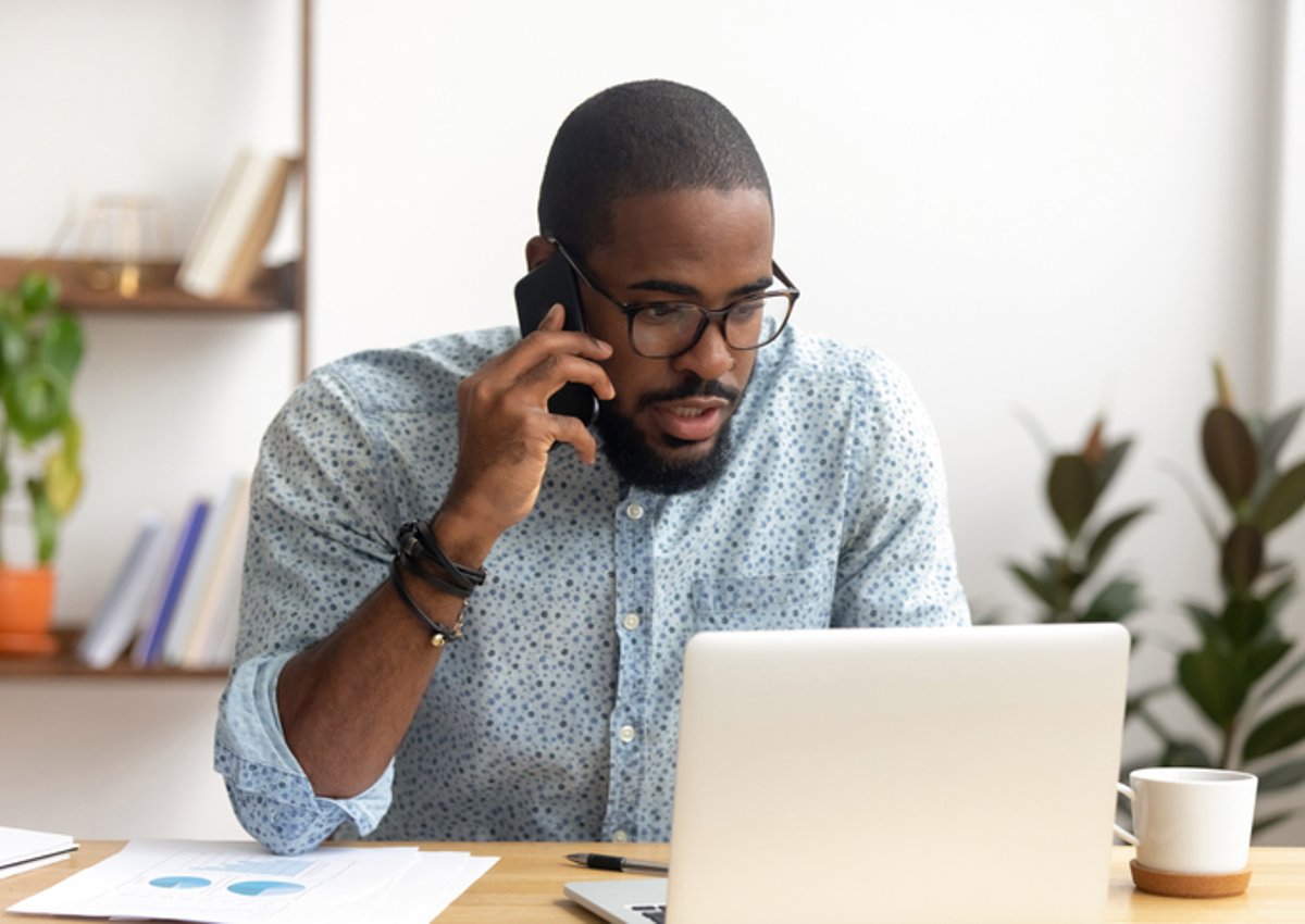 A man is sitting at his laptop at home and talking on the phone.