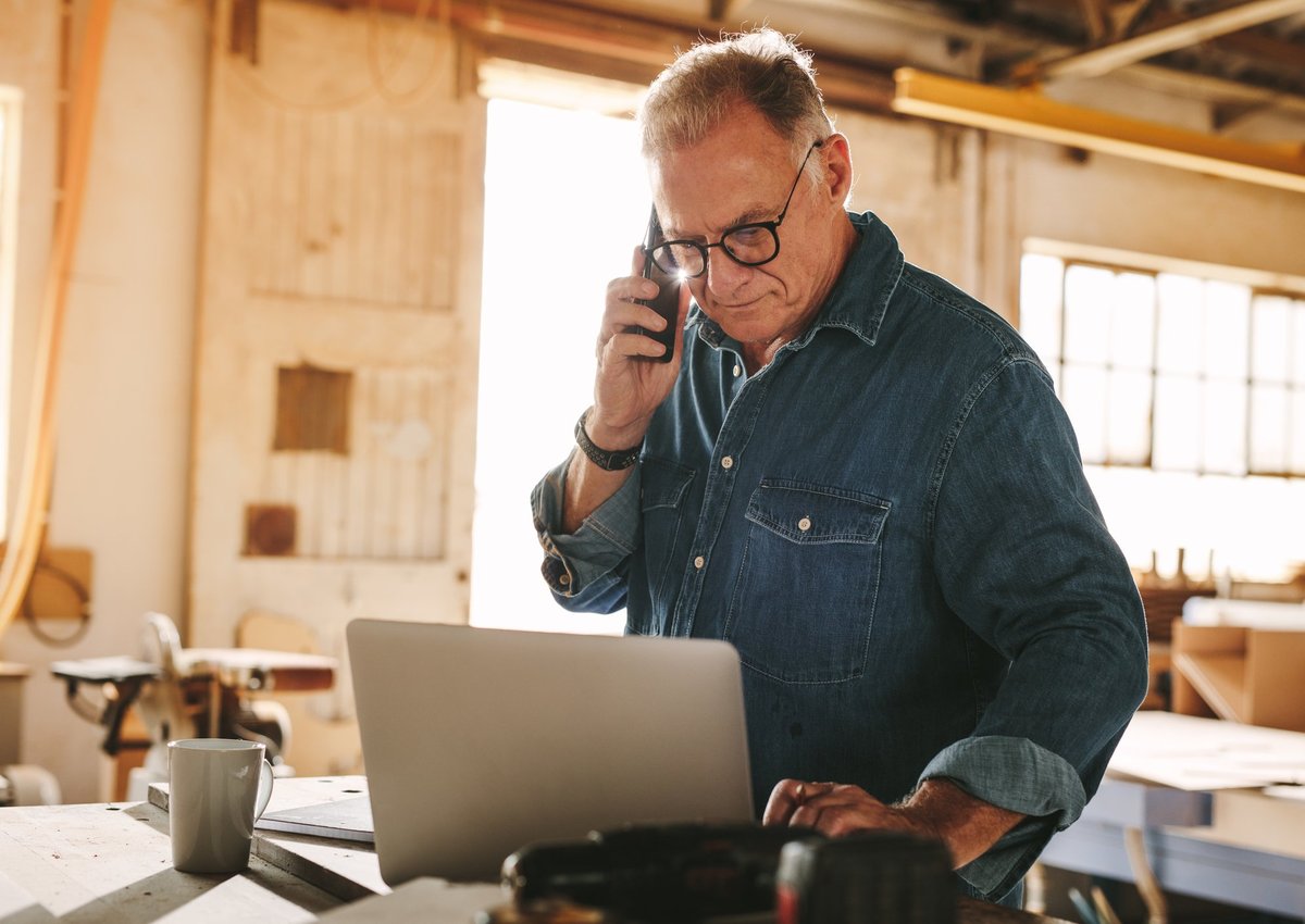 A man making a phone call while looking at his laptop in a sunny workshop.