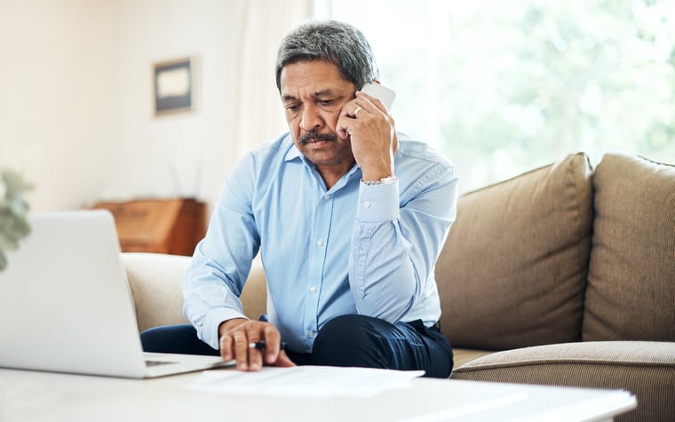 A man on a phone call while sitting on his couch with his laptop in front of him.