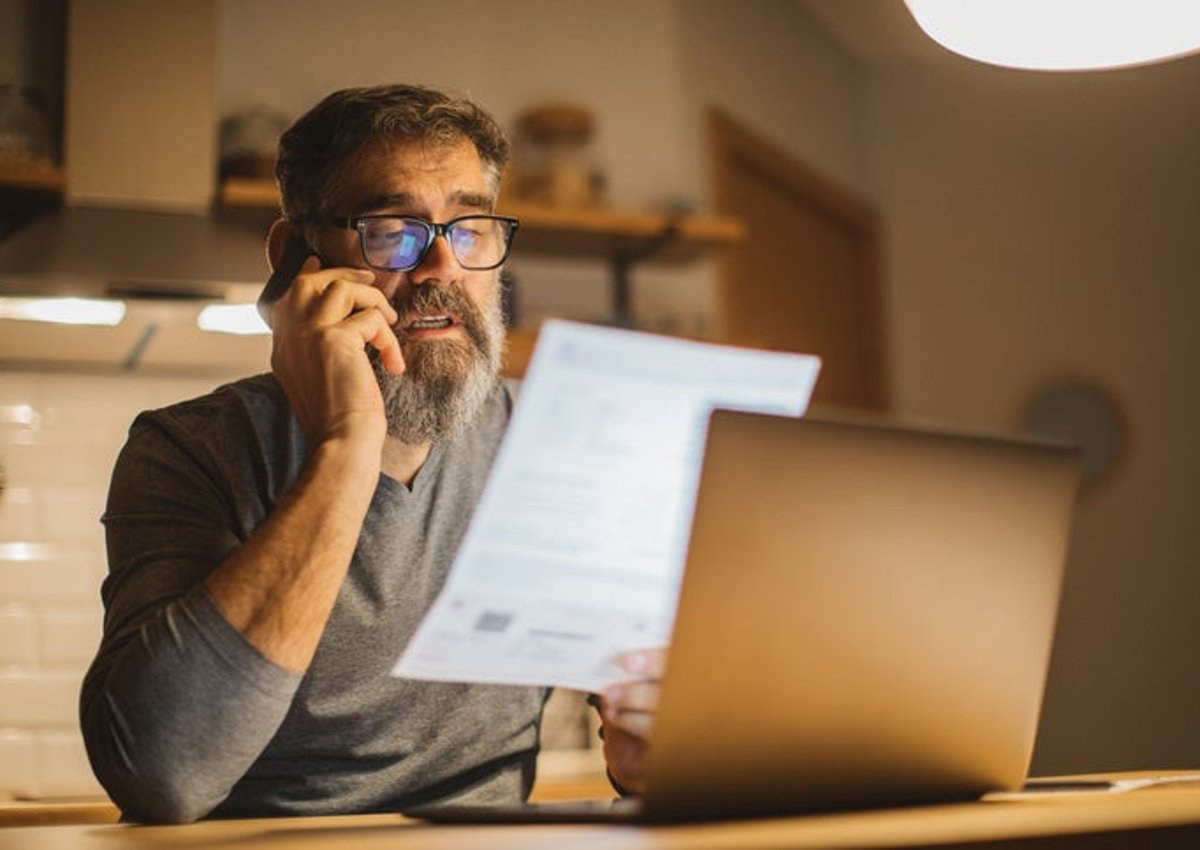 A man sitting at a desk in front of a laptop and making a phone call while holding up and reading a piece of paper.