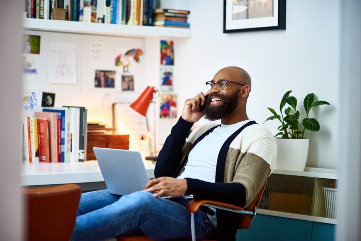 Un homme souriant au téléphone assis avec son ordinateur portable sur ses genoux tout en travaillant depuis son bureau à domicile.