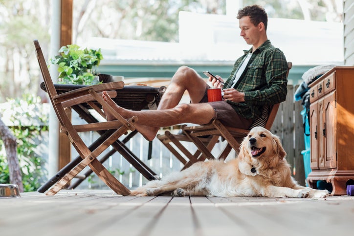 A man sitting on his sunny front porch holding a coffee and his phone with his dog lying next to him.