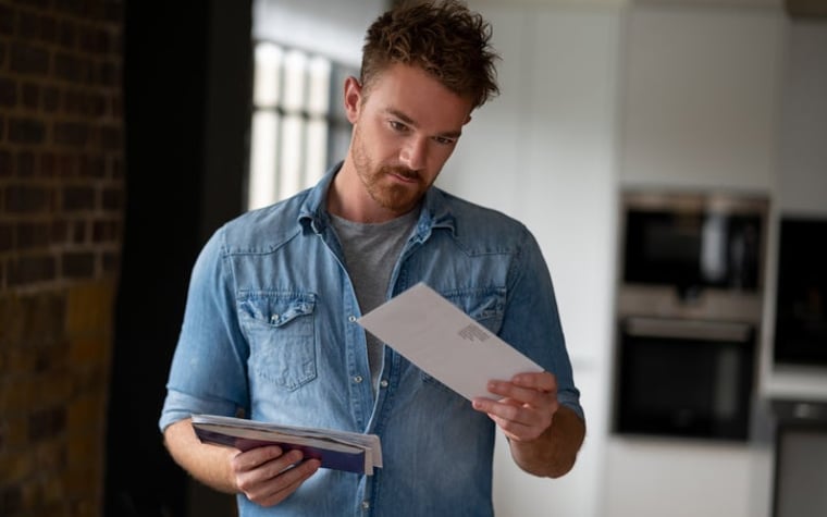 Un homme debout dans son salon et tenant une pile de courrier dans une main tout en regardant une seule enveloppe dans l'autre main.