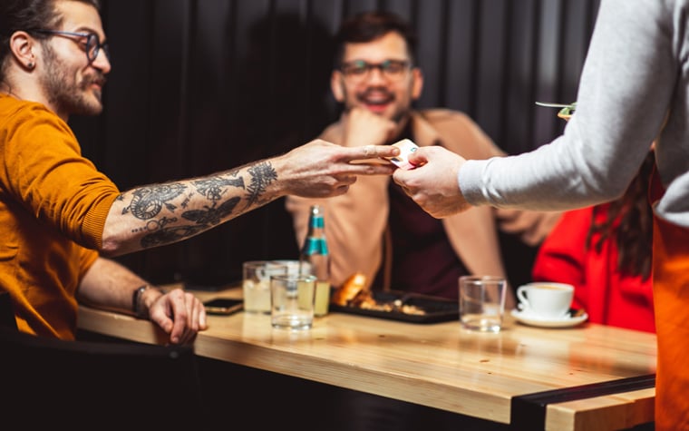 A man handing his credit card to a waiter at a restaurant while sitting at a table with his friend.