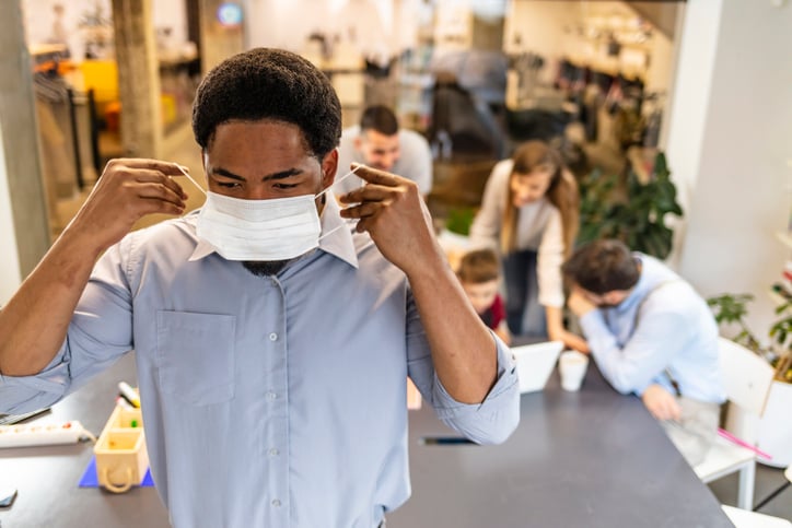 A man putting on a medical mask with his coworkers talking at a table behind him.