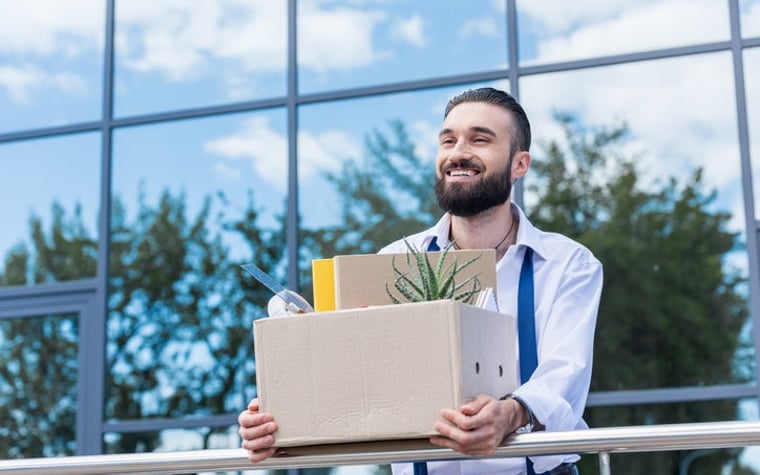 A smiling man carries a cardboard box and exits an office building.