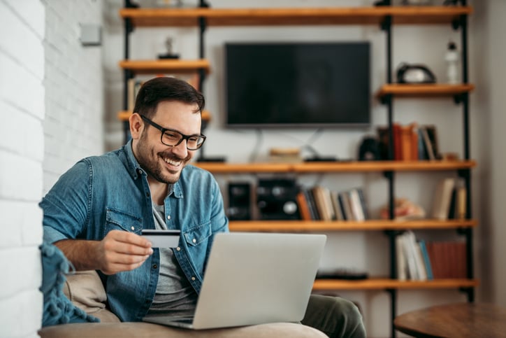 A smiling man sitting on his couch and shopping on his laptop while holding a credit card in one hand.