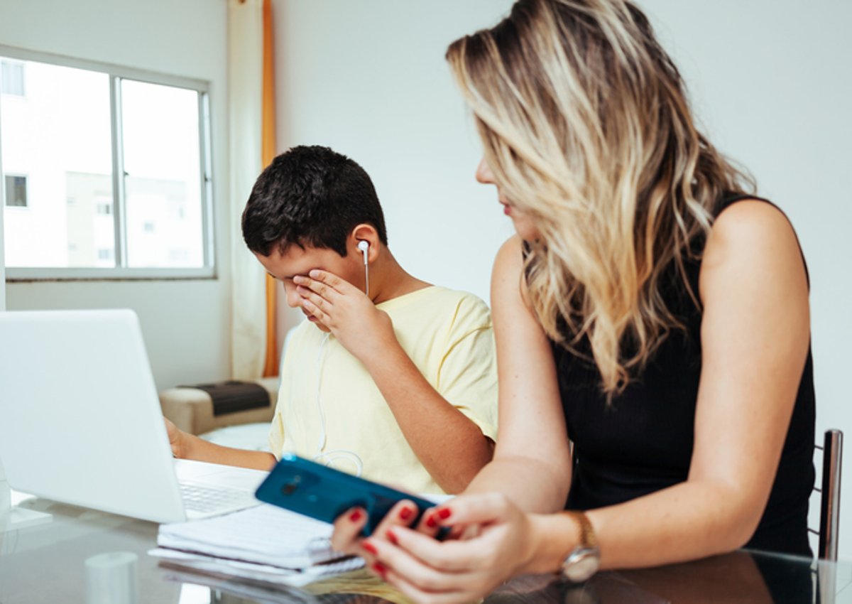 A mom sitting next to her son while he does homework and rubs his eyes at the desk in his room.