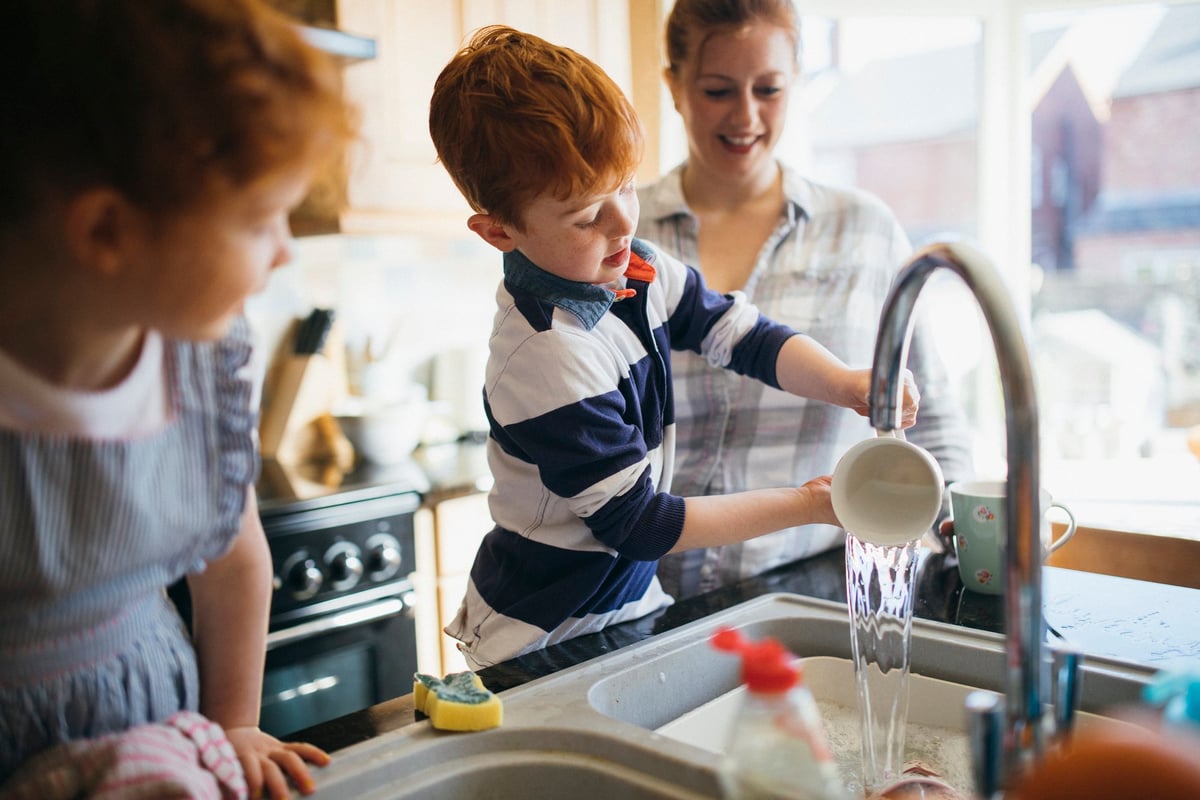 A mom and her two young children washing dishes in a bright kitchen.