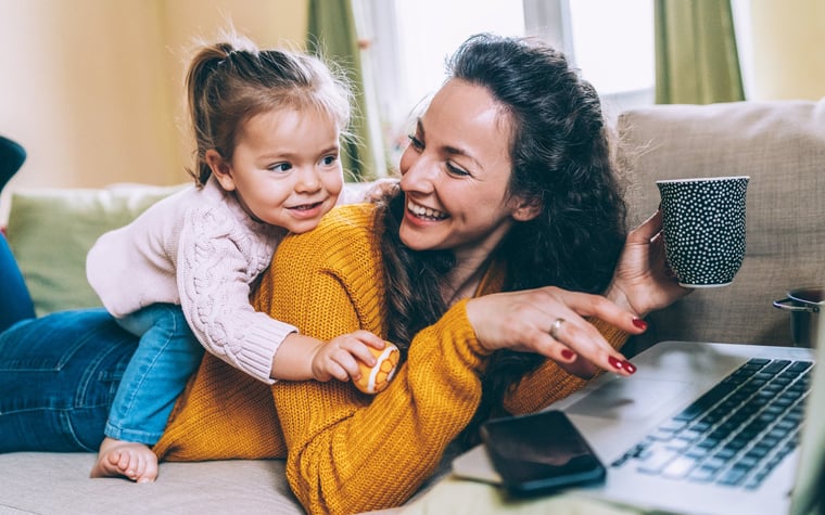 Smiling mother trying to do work on her laptop while her little daughter climbs on her back.