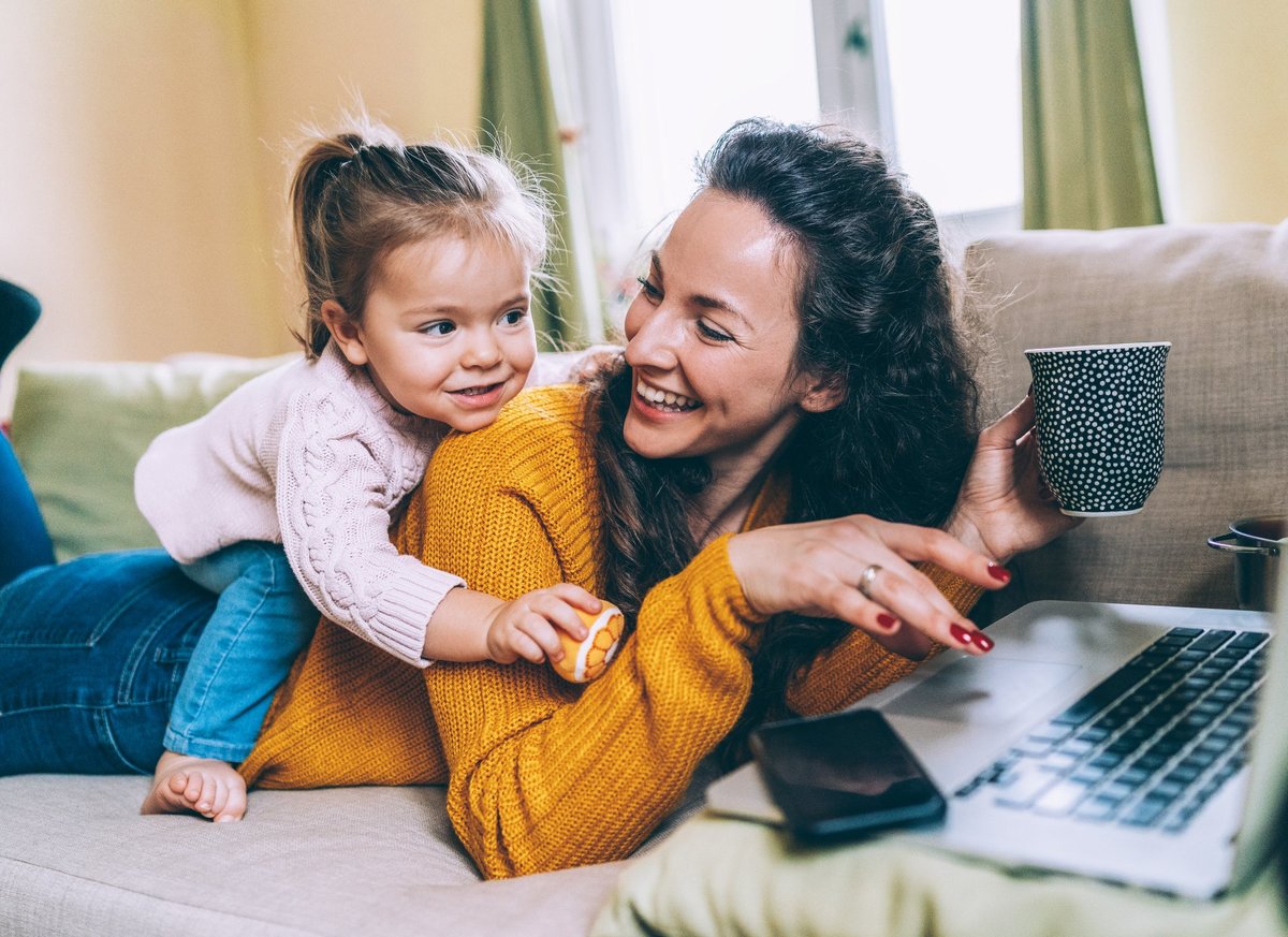 A laughing mom trying to do work on her laptop with her young daughter climbing on her back.
