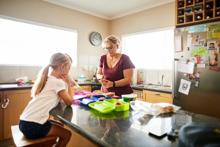 A mom standing at a kitchen island packing lunch boxes for her daughters who are sitting on stools watching.