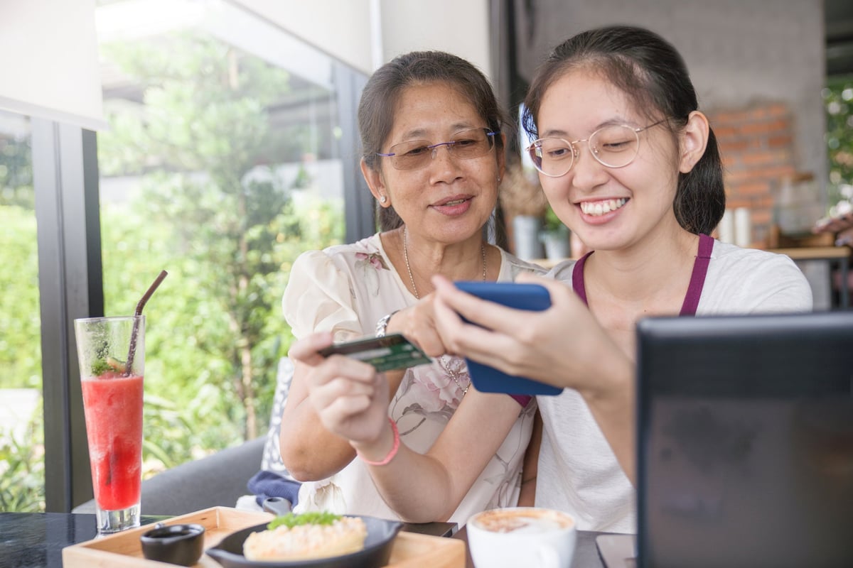 A mom sits next to her daughter at a restaurant and watches her use a credit card to pay for the meal.