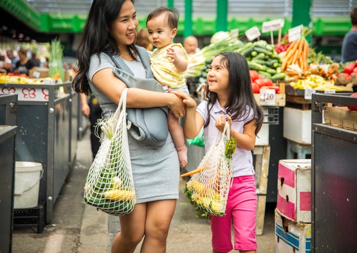 A mother holds her baby while her daughter walks next to her in the produce section of a grocery store.