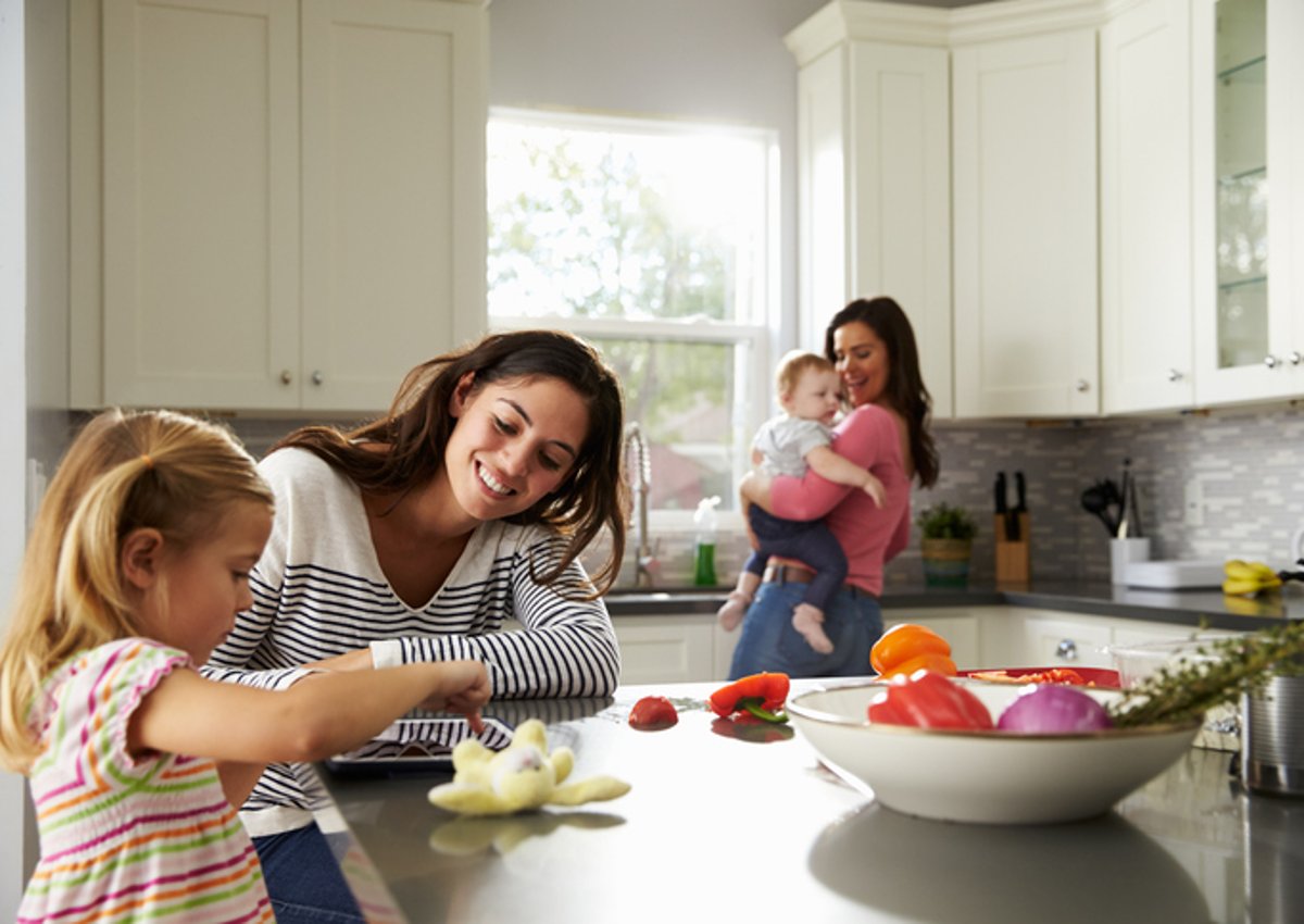 A mom playing with her daughter at a kitchen counter while the other mom dances with their baby in the background.