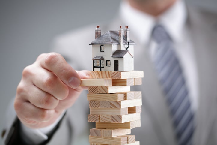 A man removes a Jenga block from a stack of blocks with a house on top.