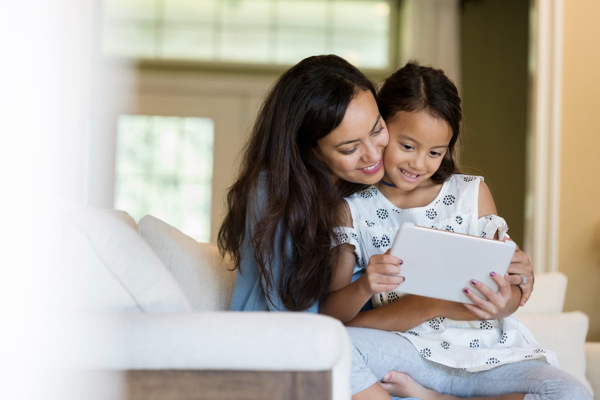 A mother and her young daughter cuddling on the couch while looking at a tablet computer.