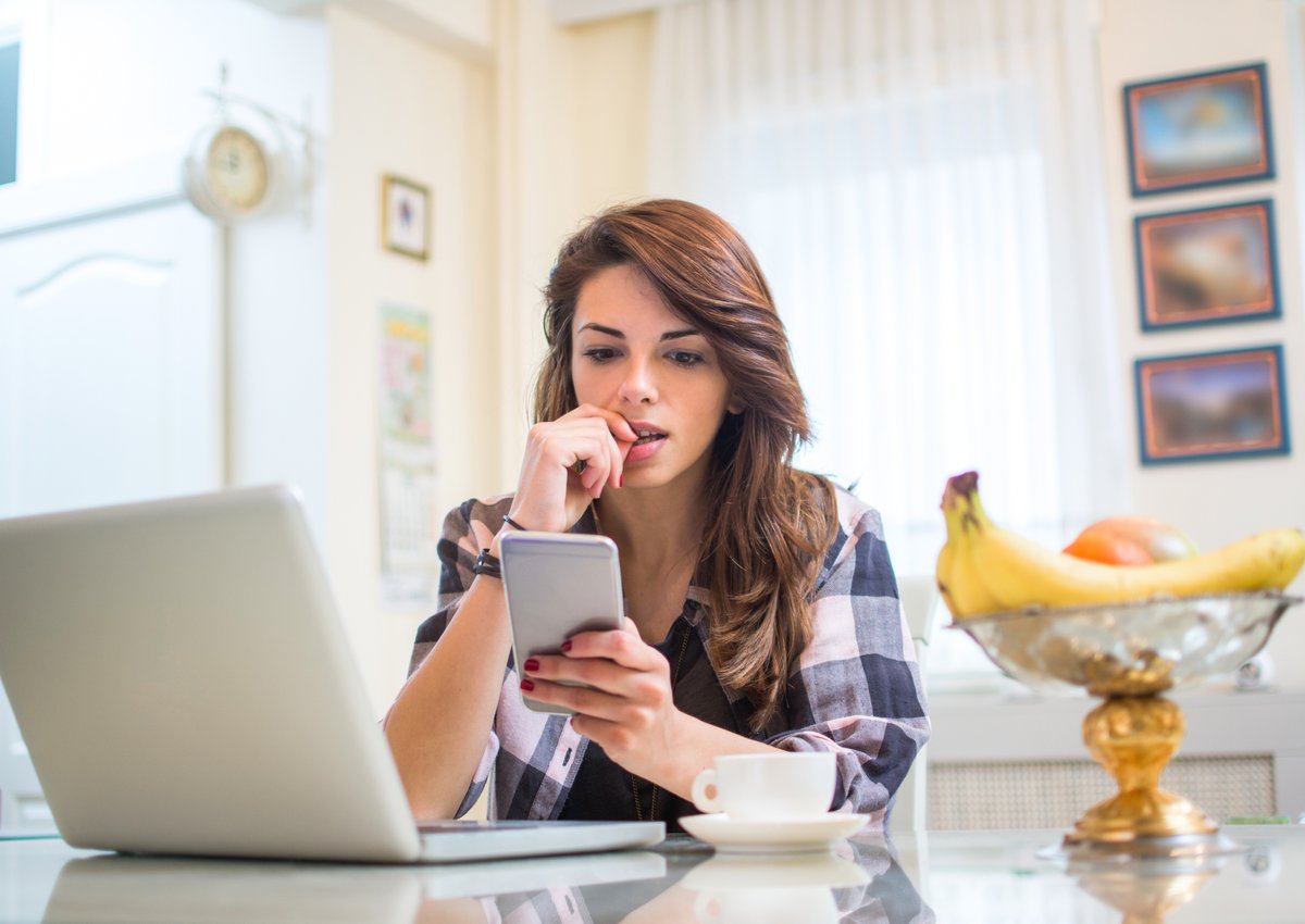 A nervous young woman biting her fingernail while sitting in her kitchen with her phone and laptop.