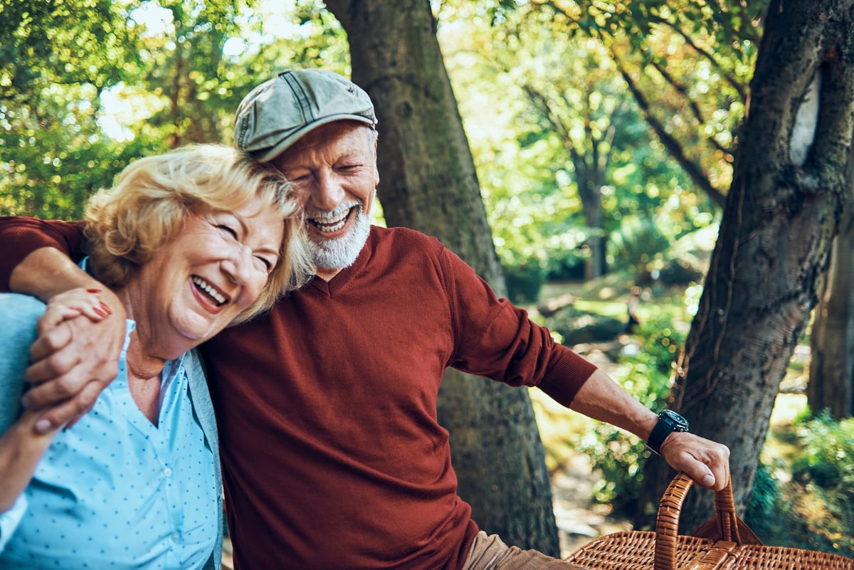 An older couple smiling with their arms around each other and holding a picnic basket in the woods.