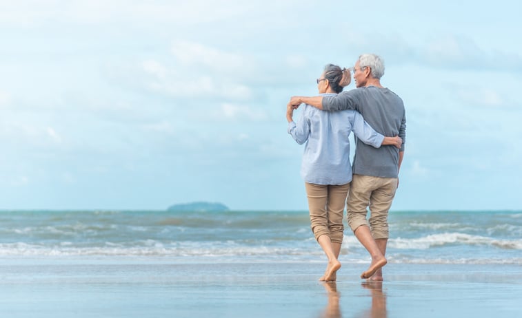 An older couple walking along the beach with their arms around each other.