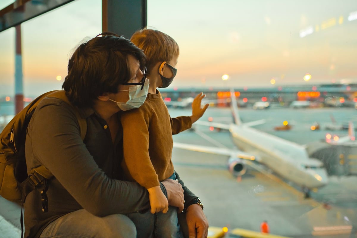 A parent and young child wearing medical masks and looking out the window of an airport.