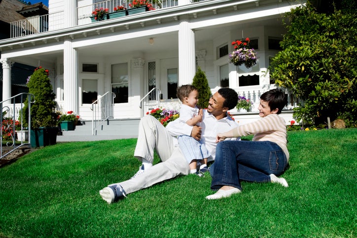 A mom and dad with their baby sitting on the sunny lawn in front of their house.