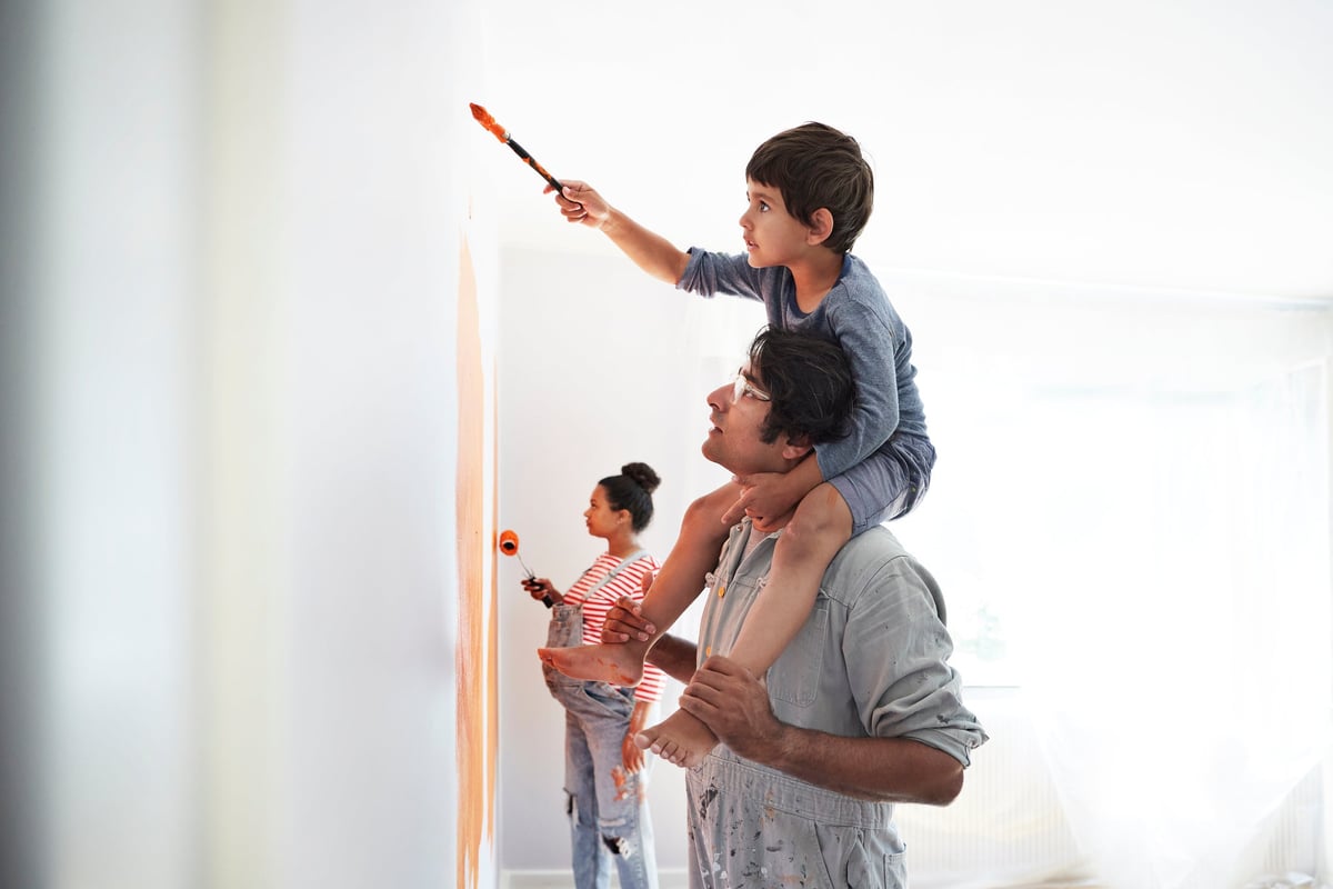 A child sitting on his parent's shoulders and painting the walls of a room while the other parent paints behind them.