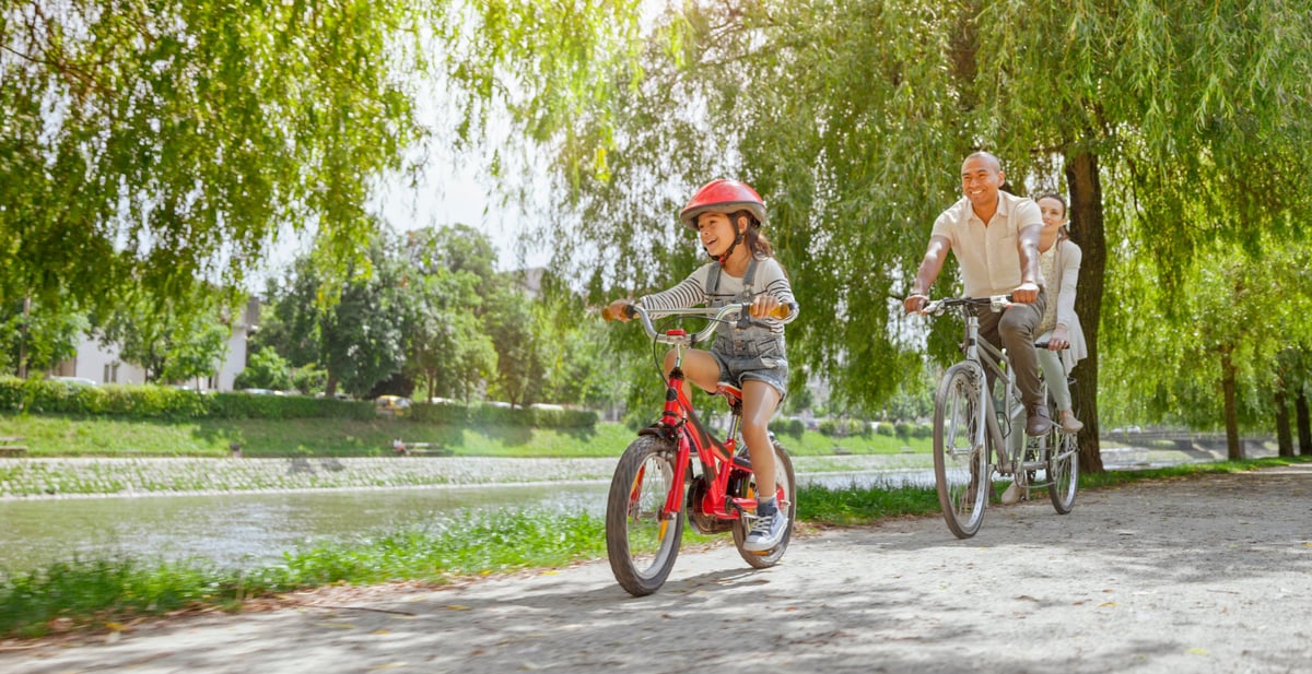 Two parents riding a tandem bike behind their young child ahead of them on a tree-lined bike path.