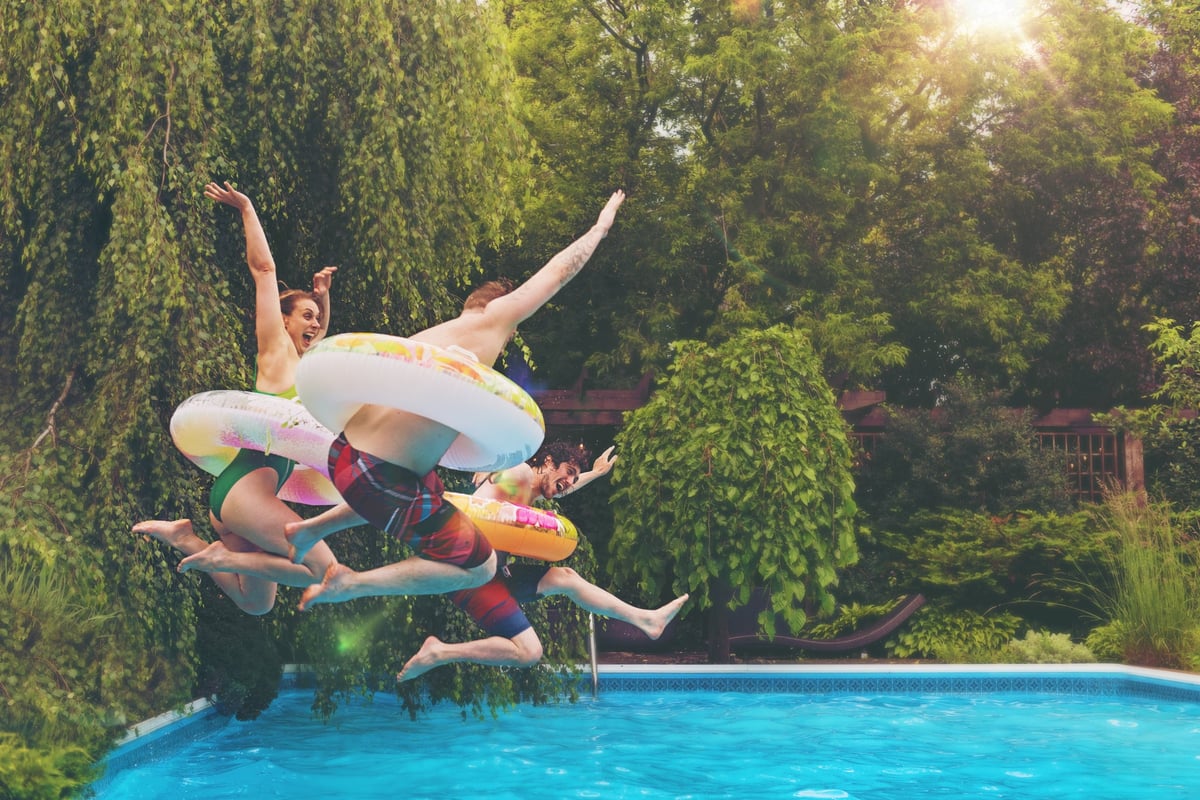 Three people wearing colorful tubes around their waists jumping into a backyard pool surrounded by trees.