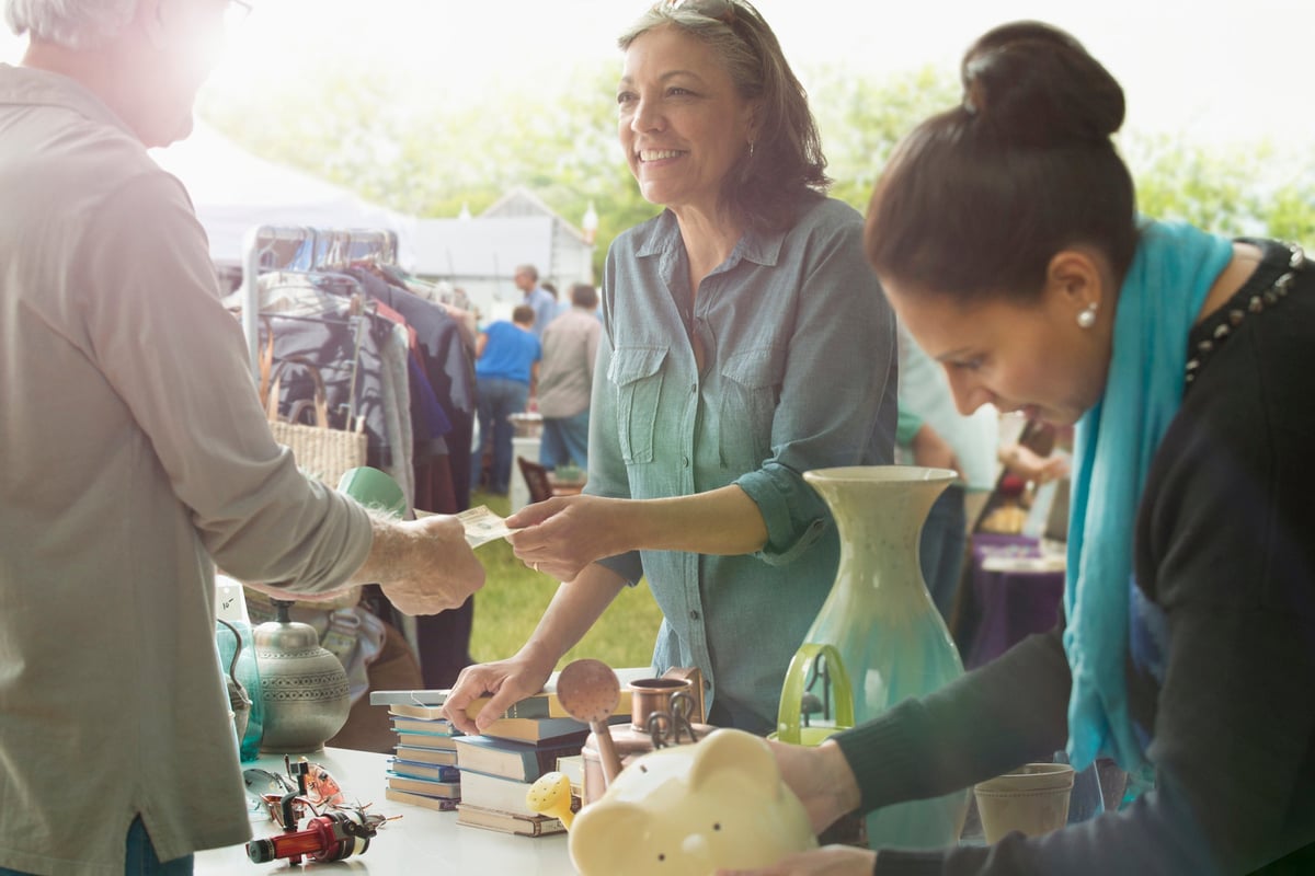 Two people looking at items at a yard sale while one hands money to the seller behind the table.
