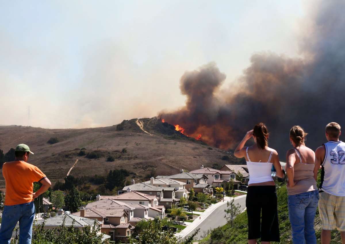 Four people watching a brush fire burn on a hill behind a street lined with houses.