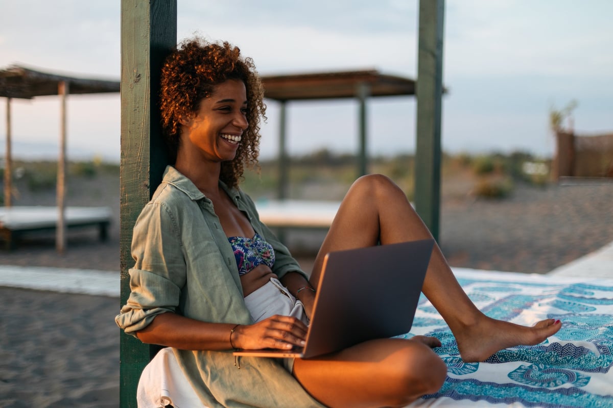 A smiling person sitting on a cabana bed on the beach while typing on a laptop.
