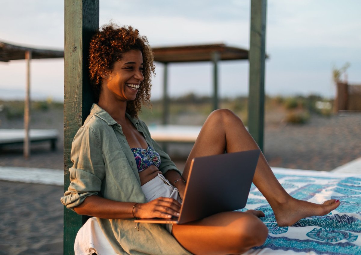 A smiling person sitting on a cabana bed on the beach while typing on a laptop.