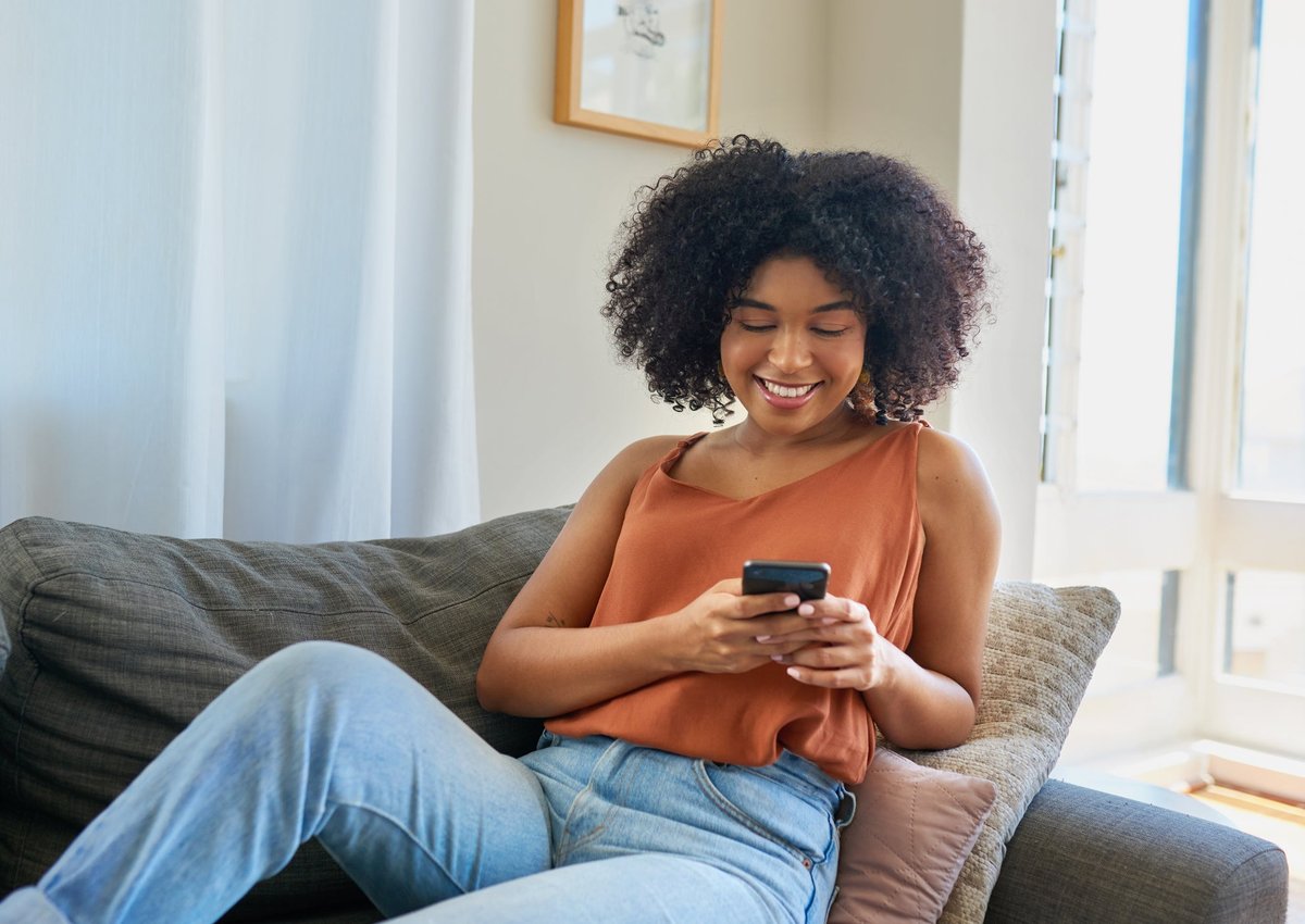 A smiling person sitting on their couch and looking at the phone in their hand.
