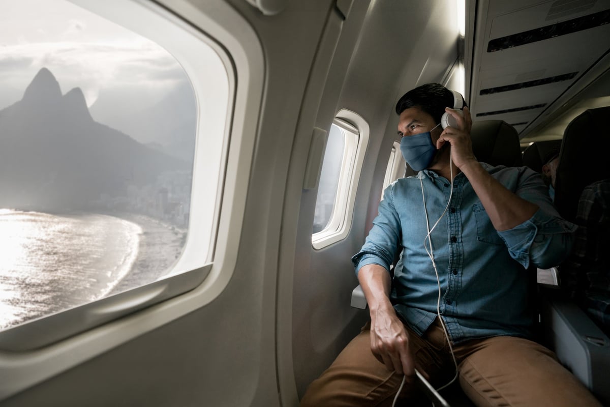 A person wearing a mask while sitting in a plane seat and looking out the window at a mountain next to the ocean.
