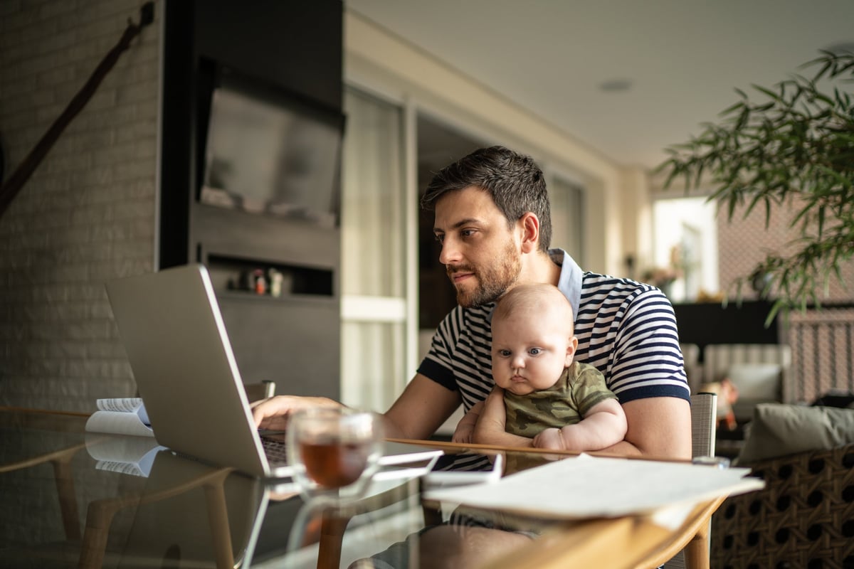 A person working on a laptop at a dining room table with a baby on their lap.
