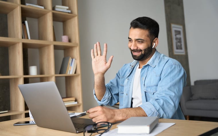 A person working at a desk at home and waving during a video call.