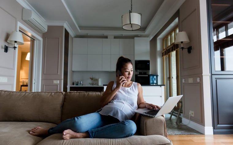 A pregnant woman sitting on her couch while searching on her computer and making a phone call.