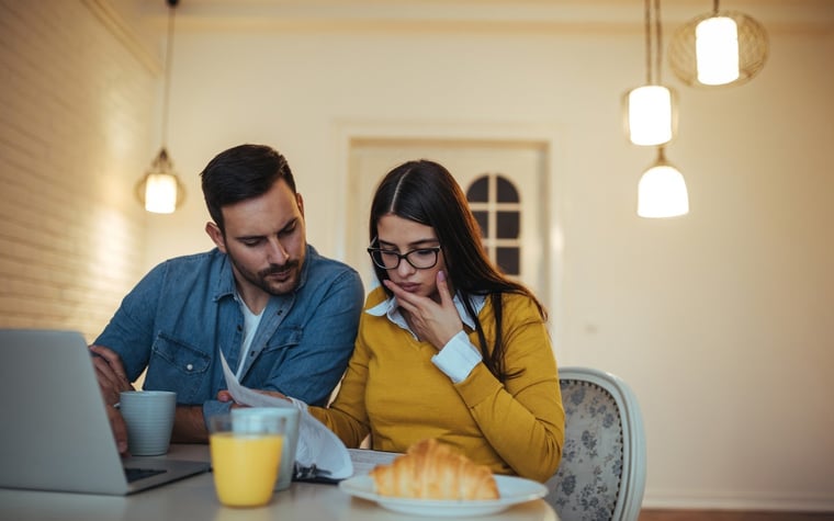 Couple eating breakfast looking at laptop and papers concerned.