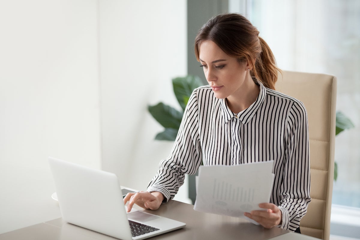 A women wearing a striped collared shirt sitting at a desk with a laptop and holding papers with charts.