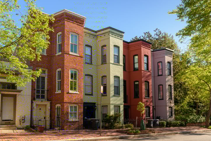 A row of colorful multistory houses in a city on a sunny street with green trees.